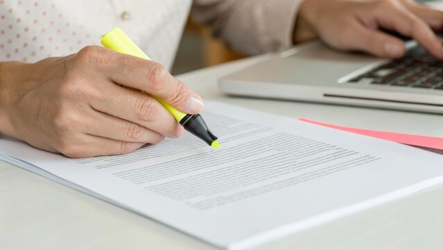 A person highlights text on a document with a yellow marker while using a laptop, suggesting multitasking or reviewing work.