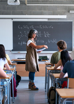 Vertical. Adult female teacher stands by the blackboard explaining a science formula to a diverse group of students in a modern classroom during a lesson.