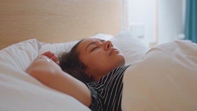 Hotel morning sunrise young woman lying in bed with soft light through curtains, gentle stretch and slow blink as guest transitions from sleep to wake, white duvet and wooden headboard frame serene