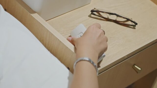 Guest dims bedside lights with remote, glasses placed beside control on nightstand, warm evening glow, finger taps dimmer button to set mood, calm restful atmosphere, minimalist hotel interior perfect