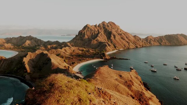Golden hour sunlight illuminating the iconic volcanic hills and tri-colored beaches of Padar Island, with traditional phinisi boats floating on the calm turquoise sea in Komodo National Park