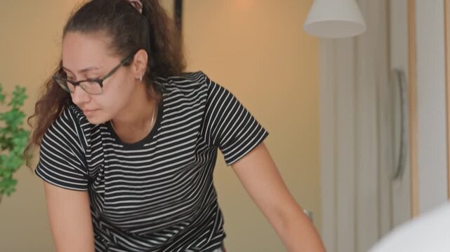 Housekeeper making hotel bed with care, hands tucking sheets and smoothing linen, adjusting pillows and mattress corner, efficient steady motions, bright hotel interior, bedside vase and lamp visible,