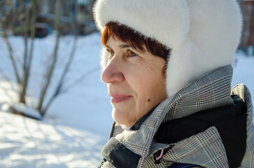 Senior woman in white fur hat and plaid coat standing outdoors in snowy winter park. Profile portrait, calm expression. Active aging, winter wellness, outdoor lifestyle, healthy retirement living © Ninaveter