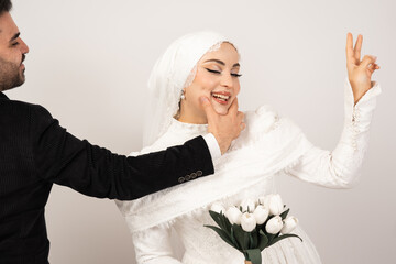 Naklejka premium Playful studio portrait of a happy bride and groom sharing a joyful moment on their wedding day. The groom gently holds the bride’s chin while she smiles and makes a peace sign gesture