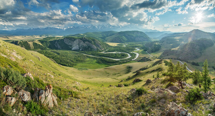 Above a picturesque mountain valley with a river, evening light, sun among the clouds