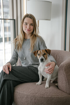 Blonde woman and dog Jack Russell terrier sitting in pink chair looking at camera. Natural light. vertical composition