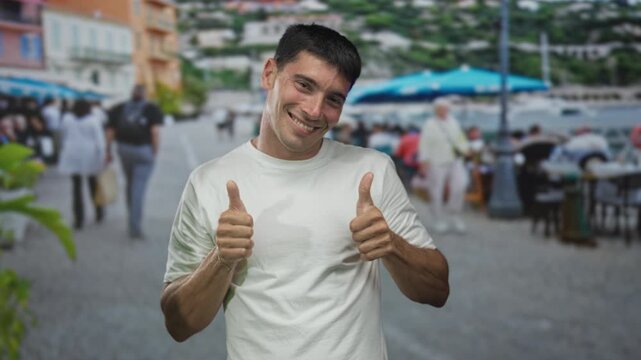 Young hispanic man wearing a white t-shirt gives double thumbs up on an outdoor restaurant terrace; optimism.