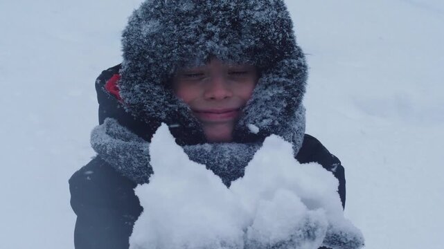 Happy boy in a warm fur hat holding a handful of fresh snow in front of him. Portrait of a child enjoying a winter walk and falling snow