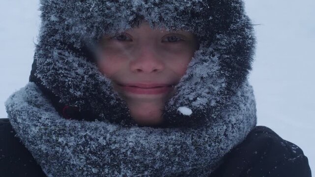 Close-up of a smiling boy in a fur ushanka hat covered in frost and snow. Portrait of a happy child during a heavy blizzard and snowfall.