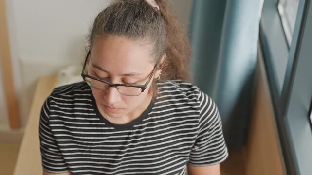 Multiracial woman guest hotel tasting snack by window food blogger leans over table bites cookie sips green tea glasses focused expression striped shirt natural daylight boutique hotel corner content
