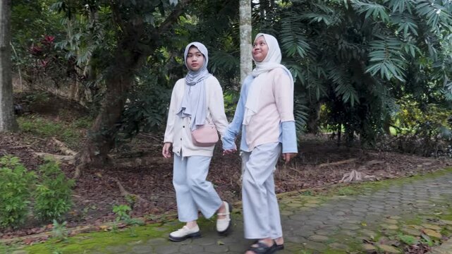 Two young Asian Muslim sisters walking through a lush forest park, chatting and enjoying nature together under the morning sun