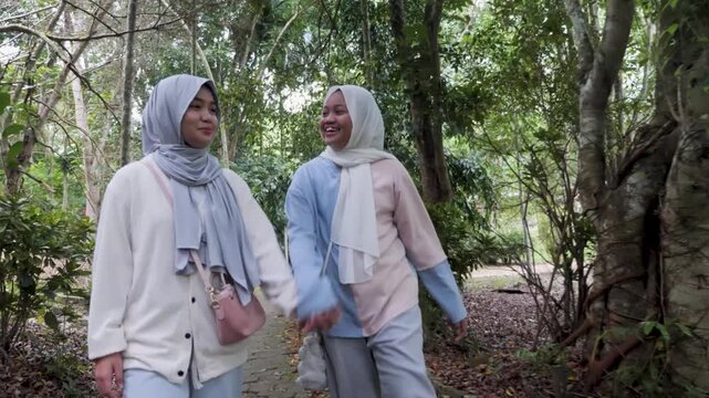 Two young Asian Muslim sisters walking through a lush forest park, chatting and enjoying nature together under the morning sun