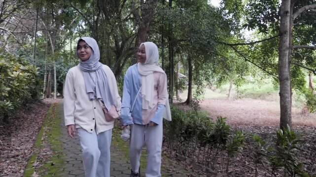 Two young Asian Muslim sisters walking through a lush forest park, chatting and enjoying nature together under the morning sun