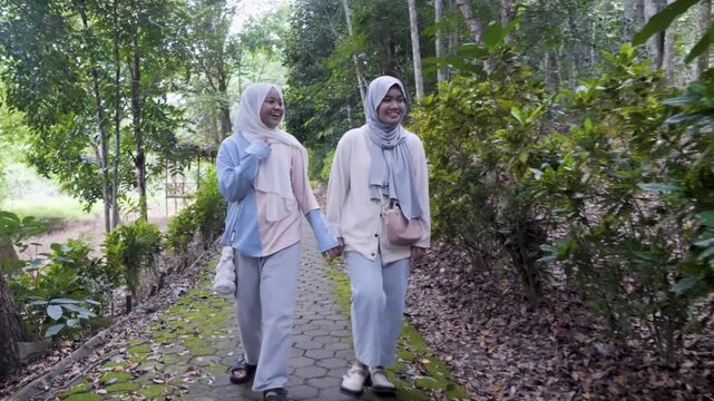 Two young Asian Muslim sisters walking through a lush forest park, chatting and enjoying nature together under the morning sun