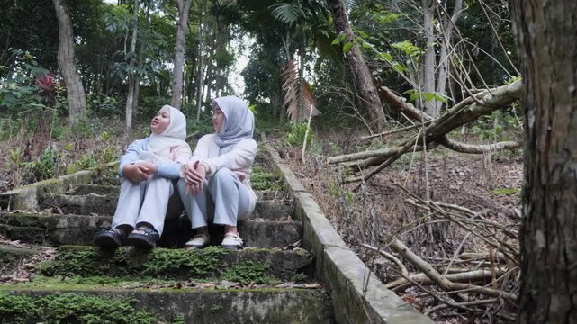 Two young Asian Muslim sisters sitting on forest park stairs, chatting and enjoying nature together under the morning sun
