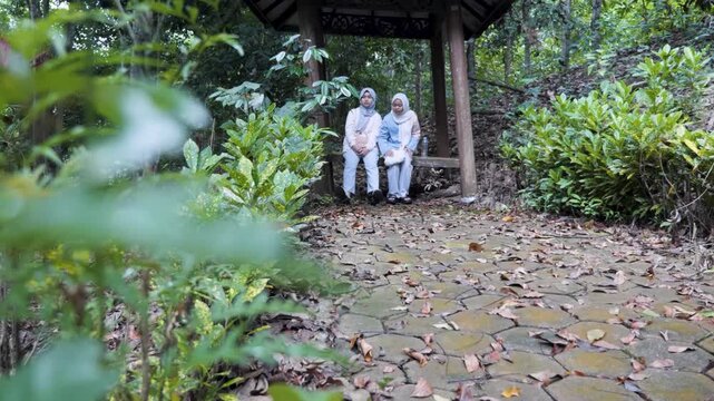 Two young Asian Muslim girls sitting and resting in a wooden gazebo, chatting and enjoying nature together under the morning sun in a forest park
