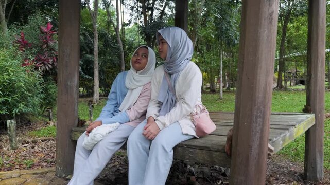 Two young Asian Muslim girls sitting and resting in a wooden gazebo, chatting and enjoying nature together under the morning sun in a forest park