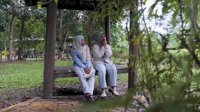 Two young Asian Muslim girls sitting and resting in a wooden gazebo, chatting and enjoying nature together under the morning sun in a forest park