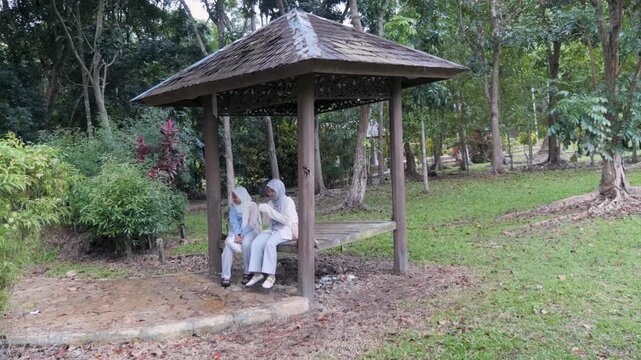 Two young Asian Muslim girls sitting and resting in a wooden gazebo, chatting and enjoying nature together under the morning sun in a forest park
