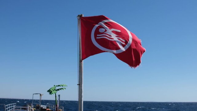 Red warning flag displaying swimming prohibition symbol waves in the breeze against a bright blue sky over the ocean near a dock with palm tree