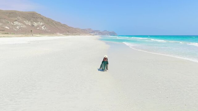 Aerial view of a young woman walking on a white sand beach along the turquoise ocean near mountains on Socotra Island, Yemen.