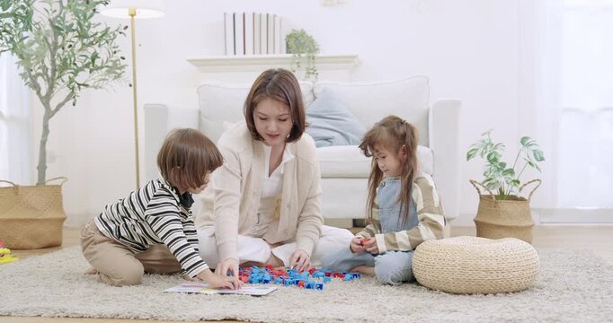 Asian single mother guides son, daughter teamwork building toy bricks together on soft carpet, living room. Kids creative play supports early childhood development, counting practice, quality time.