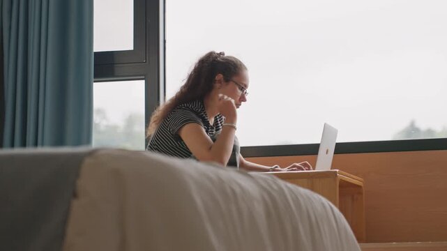 Female author typing at hotel bedside table. Woman diligently drafting notes on her laptop in calm hotel room. Focused woman writes on her laptop in cozy hotel setting with muted decor