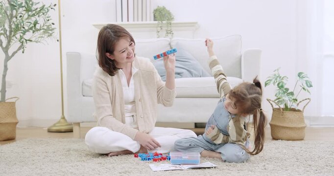Asian single mother laughing, holding letter block name, prompting preschool daughter raise arm and respond, practicing alphabet spelling, boosting confidence, weekend family bonding livingroom.