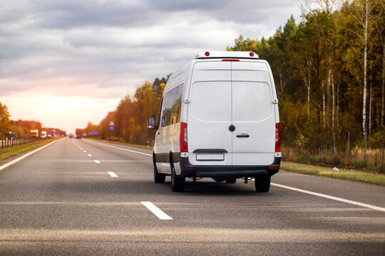 White cargo van driving on highway at sunset, autumn trees line road, delivery vehicle traveling on asphalt route during golden hour with cloudy sky
