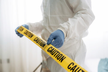 Forensic expert in protective gear holds yellow caution tape in a well-lit indoor setting, indicating a crime scene investigation with blurred background elements