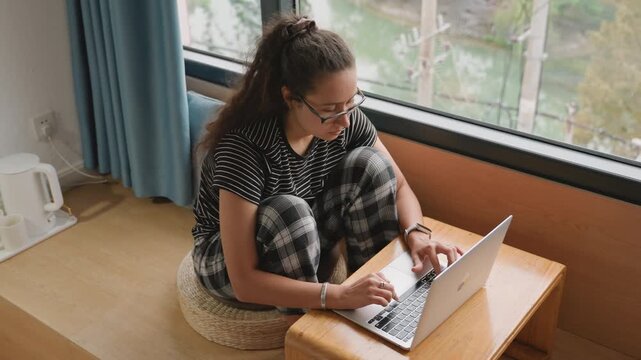 Relaxed woman using computer by window. Casual scene of female worker at hotel with laptop near window. Woman working comfortably with her laptop at hotel window during leisure time