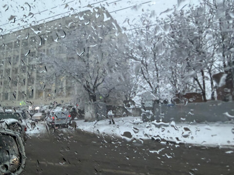 Raindrops on car window during snowy day in city. Blurry urban winter view from inside vehicle. Cold weather concept with melted sleet and snow covered trees in gloomy winter season.