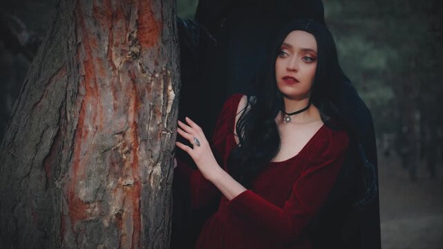 Fairytale Gothic scared girl in arms of death angel, monster nightmare stands behind scares touches victim in night dark forest trees fog night. Fantasy Woman red old style dress. Black hair sexy face