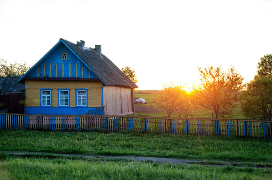 Rural house with vibrant blue and yellow facade and old wooden fence at sunset. Green field of dandelions in foreground. Peaceful countryside landscape during golden hour.