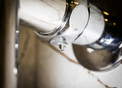 Water drop forming and falling from polished chrome towel warmer surface in modern bathroom, close-up view of plumbing leak and mineral deposits showing maintenance issue