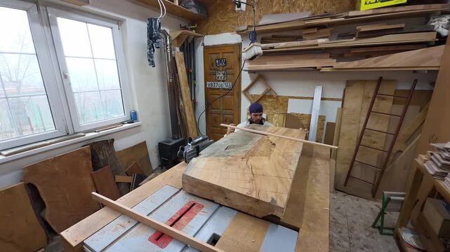 Authentic Male Artisan Carpenter Evaluating Live Edge Timber Slab Flatness with Straight Edge in Messy Carpentry Workshop Wide Angle Handheld Documentary Shot