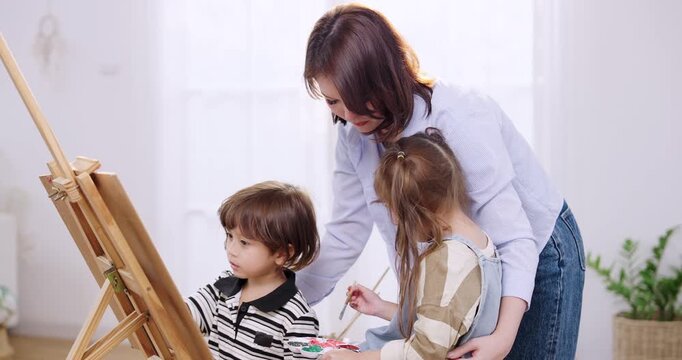 Asian single mother leans close, guides son watching sister mix paint on palette, homeschooling preschool learning game with toy blocks sparks counting talk, family relationship, indoor living room