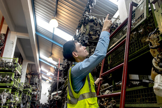 Young Asian male engineer worker with safety vest inspecting at auto spare parts warehouse. Industrial labor man checking and counting product while climbing the ladder surrounded by many engine parts