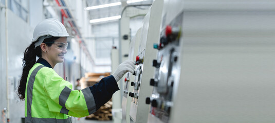 Plakat Caucasian female factory technician wearing safety helmet, gloves, goggles and reflective vest operating industrial machine using control panel in cardboard packaging factory production line.