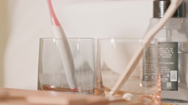 Hotel bathroom vanity with toothbrushes and drinking glasses on wooden tray, caucasian hand placing pink electric toothbrush beside glass, warm natural light, tidy amenity bottles, closeup texture