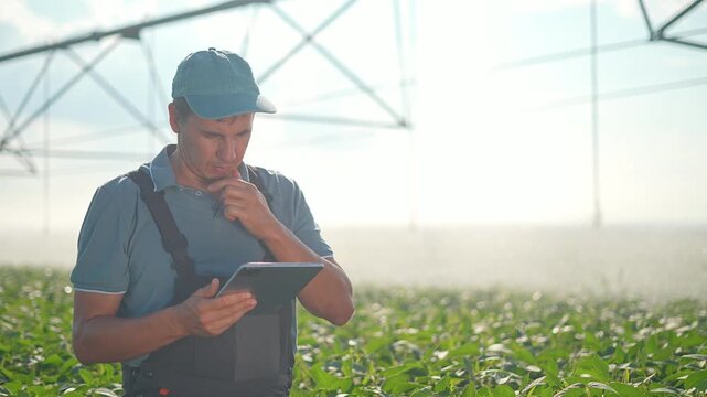 Farmer uses digital tablet in green field. Agriculture worker checks smart farming data on tablet. Farmer touches screen near irrigation. Technology helps agriculture. Man works in crop field.