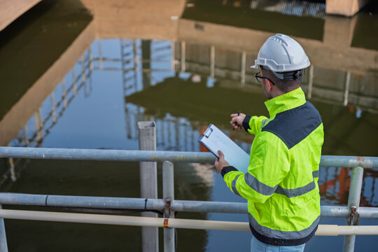 Professional Asian engineer inspecting water quality at a wastewater treatment plant. He is carefully recording maintenance data on a clipboard during an industrial site safety survey.