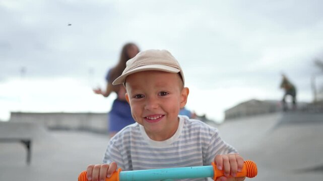 Excited boy rides scooter at skate park while mother watches nearby. Happy child learns scooting outdoors. Boy holds scooter handlebar laughing. Mother supports excited child riding scooter.