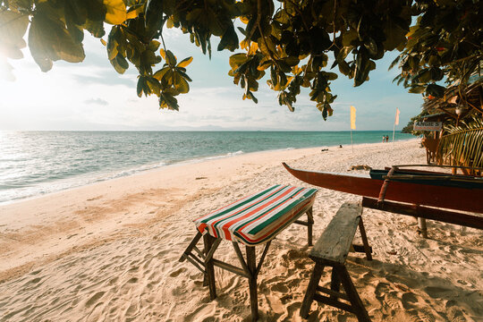 View of sun-kissed sand, turquoise waters, and a rustic bench under the shade of leafy trees, inviting relaxation on a tropical beach, Badian, Central Visayas, Philippines.