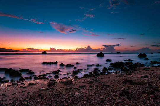 View of a tranquil beach with rocks scattered in the foreground and a vibrant sunset painting the sky with hues of blue, orange, and pink, Mati, Davao Region, Philippines.