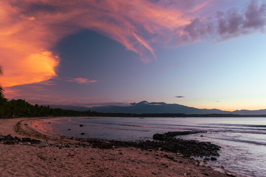 View of a tranquil beach where the soft sand meets gentle waves under a sky ablaze with fiery orange and fading purple hues, Mati, Davao Region, Philippines.