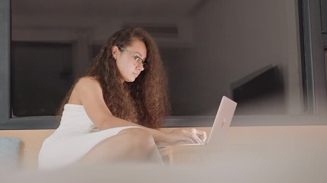 Female student studying. Lady diligently revising notes during evening with cityscape view. Female scholar intensely focused on her laptop at night in serene hotel setting with city lights