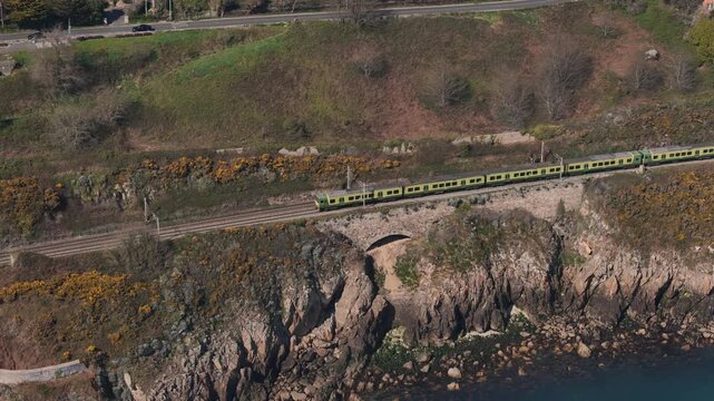 Drone footage capturing the DART train gliding along the Dalkey coastline in Dublin, with waves crashing below and cliffs rising beside the tracks
