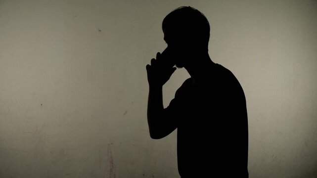 Anonymous person holds phone near face while standing in front of a blank wall in a simple room during evening hours