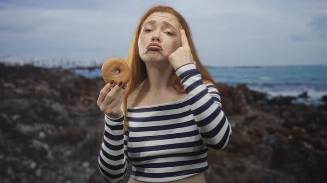 Redhead woman holding a glazed donut and pinching her cheek with index finger at rocky beach shoreline wearing a striped crop top; temptation.